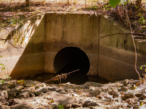 Concrete drainage culvert pipe outlet in a dry rocky ditch surrounded by autumn trees and foliage