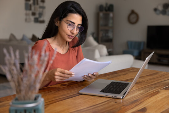 Thoughtful document review. Busy young woman freelance accountant lawyer analyst engaged in paperwork at cozy home office study marketing report research papers compare data with information on laptop