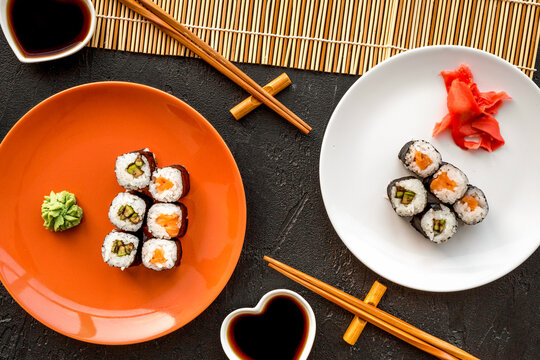 Sushi roll with salmon and avocado on plate with soy sauce, chopstick, wasabi on mat on black background top view