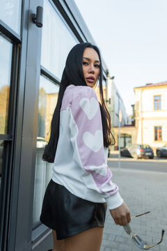 Stylish Asian woman with long dark hair looking back while holding sunglasses, wearing a white sweater with pink heart-patterned sleeves and black leather shorts outdoors by a glass building