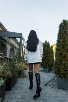 Back view low angle shot of a young woman with long dark hair, wearing a white sweater, black shorts, and high leather boots, walking down a paved street near green trees and cafe.