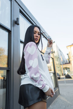 shot of a beautiful Asian woman looking back over her shoulder, holding sunglasses, wearing a pink-and-white sweater and leather shorts outdoors by a glass building.