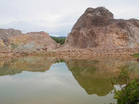 Rugged brown rock formations at an abandoned quarry reflected in a calm water pool under a cloudy sky, creating a natural landscape with exposed rocky surfaces and surrounding greenery.