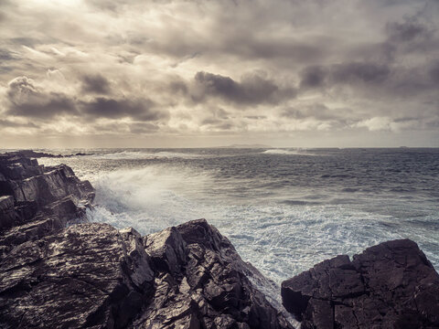 Stormy ocean waves crashes against rugged, weathered rocks. Grey clouds loom over distant, mist shrouded hills. Waves churn with force, creating white foam and spray. West coast of Ireland.