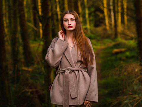 Woman in cozy coat stands among tall trees. She gazes calmly with soft, thoughtful expression. Forest floor covered with moss and fallen leaves. Perfect for fashion, travel, or nature-themed projects