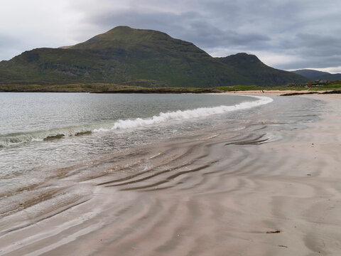 Peaceful coastal view with calm waves, wide sandy shore, and distant green hills under cloudy skies
