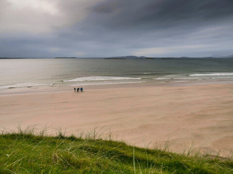 Two small figures walk along a quiet, sandy beach. Dark clouds loom overhead as gentle waves roll in. Distant islands and rocky outcrops line the horizon