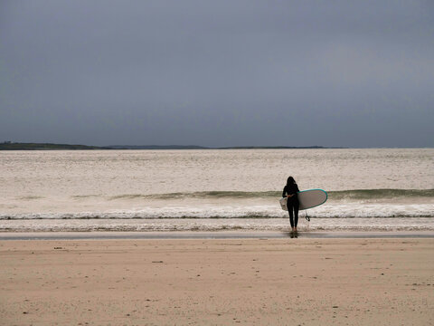 Isolated surfer carries board into sea under overcast skies