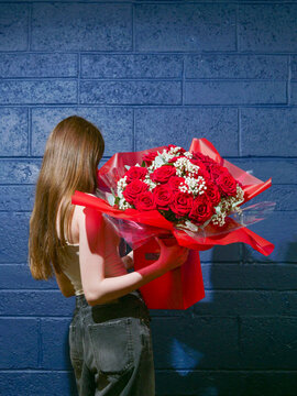 Woman holds vibrant red roses bouquet against blue brick wall. She stands with back turned, suggesting anticipation or surprise. Perfect for Valentine's Day, anniversaries, or romantic gestures