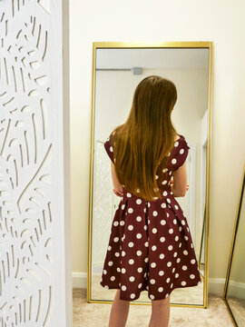 Young girl with long hair stands before tall, golden-framed mirrors. She wears a polka-dot dress, examining her fashion stylish outfit. Her posture suggests quiet curiosity and self-discovery