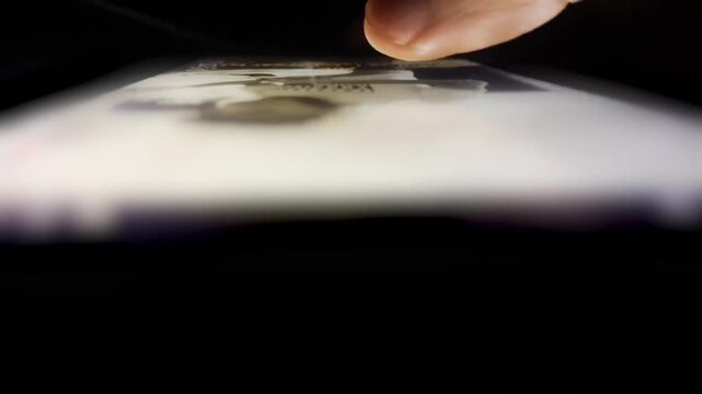 Scrolling On Social Media at Night Concept. Extreme Close-Up From A Low Angle Of A Finger Swiping Across An Illuminated Smartphone Screen In Dark 