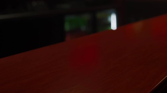 A wooden bar top sits in a dimly lit room. Soft reflections from the light create a warm atmosphere. The area appears inviting, with various blurred objects in the background.