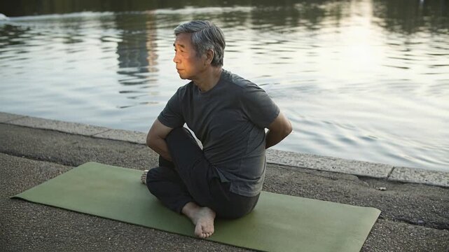 Sitting senior Asian man grey tee threading riverside, sun urging stretch on green mat, white swan