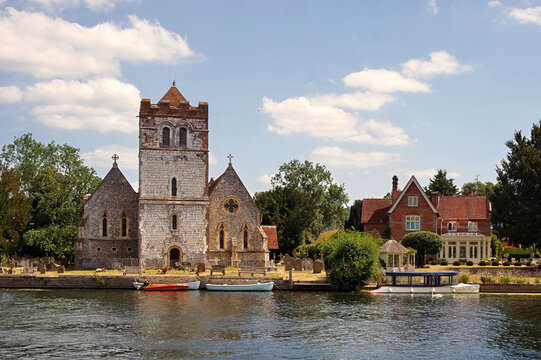 Scenic view of All Saints Church and riverside houses along the River Thames in the historic village of Bisham, Buckinghamshire, England, under a bright blue summer sky with scattered clouds.