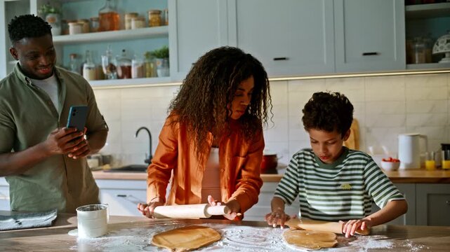 Parents son baking cookies together. Family rolling dough kitchen, dad filming.