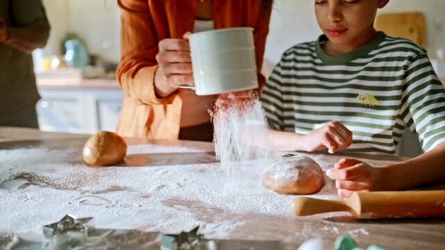 Boy rolling dough with mom. African american woman sifting flour home kitchen.