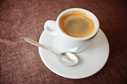 White ceramic cup filled with hot espresso coffee with crema layer on saucer with metal spoon on brown textured fabric tablecloth