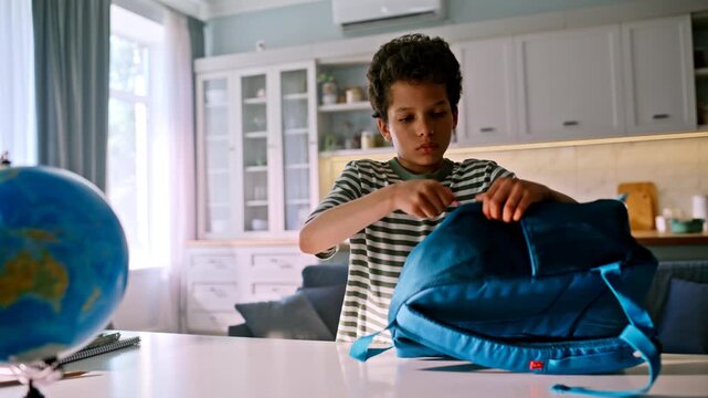 Schoolboy closing backpack zipper kitchen table. Mixed race kid leaving home.
