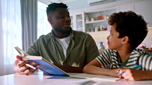 Black father son laughing over book home kitchen. Boy doing homework notebook.