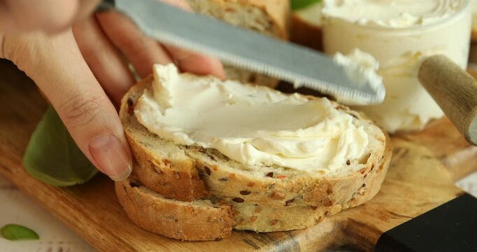 Woman spreading cream cheese onto bread slice with knife at white wooden table, closeup