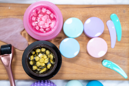 Top view of two open jars with pink and yellow capsules in clear gel beside round swatches of pastel shades and turquoise smears on a wooden board, cosmetic ingredient marketing.