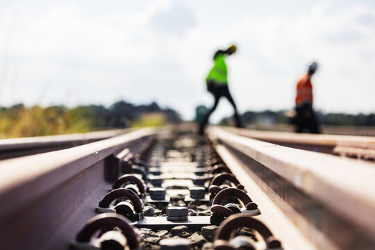 Low angle close-up of steel railway rails secured by fastening clips on concrete sleepers, emphasizing track support and structural stability, with two engineers blurred in the background.