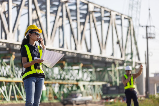 An engineer wearing a yellow hard hat and reflective safety vest reviews construction plans at a railway bridge site, with a steel truss structure in the background.