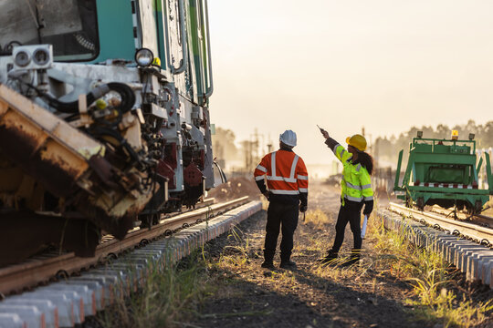 Two engineers wearing hard hats and high visibility safety clothing inspect a railway construction site beside heavy machinery. One points along the tracks while coordinating work in warm evening ligh