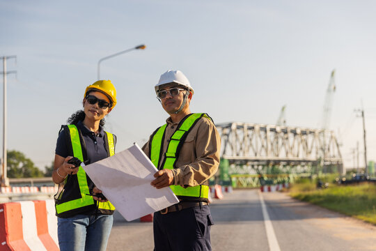 Two engineers wearing hard hats and reflective safety vests hold construction plans at a railway overpass project. One carries a handheld radio, with a steel truss bridge under construction 