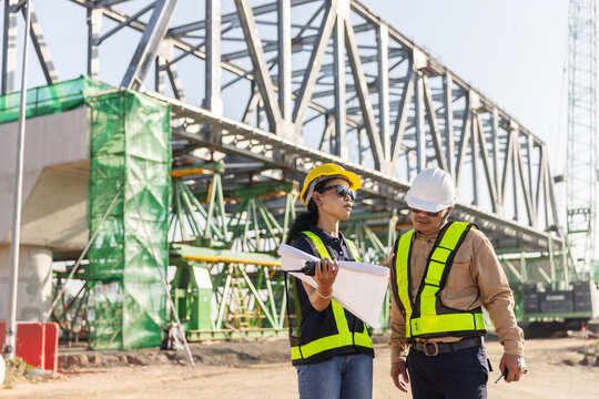 Two engineers wearing hard hats and reflective safety vests hold construction plans at a railway overpass project. One carries a handheld radio, with a steel truss bridge under construction 