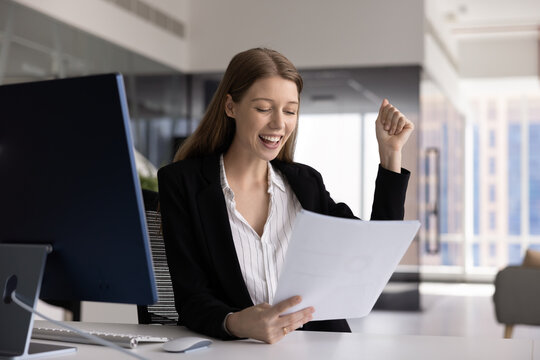 Young woman accountant or marketing manager celebrating positive audit results successful sales statistics revenue growth costs efficiency or budget surplus reflected in paper reports at office desk