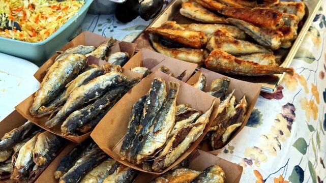 Small fried fish fill paper trays on a food display table, with larger fish portions, shredded cabbage and carrot salad, and browned patties visible alongside.