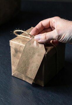 Close up of person holding blank paper tag on rustic brown paper gift box tied with twine string on black background
