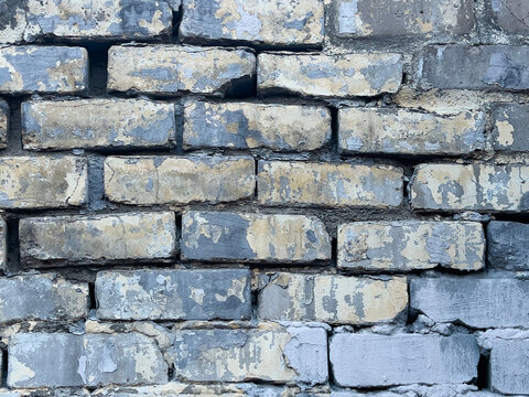 Weathered brick wall with peeling pale yellow and gray paint showing mortar cracks and textured masonry surface under soft daylight highlighting rough edges, close-up detail