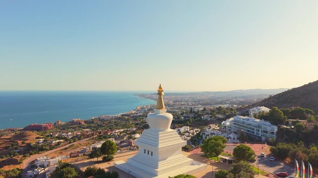 A drone rises past the golden spire of the Benalmadena Stupa to reveal hillside villas with pools and the curving Costa del Sol coastline in the evening light.