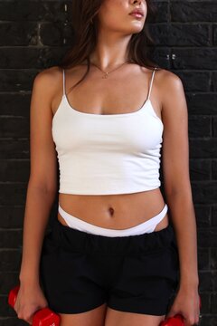 Slim fit young woman in blank white crop top and black shorts holding red dumbbells against dark brick wall, t-shirt and apparel mockup