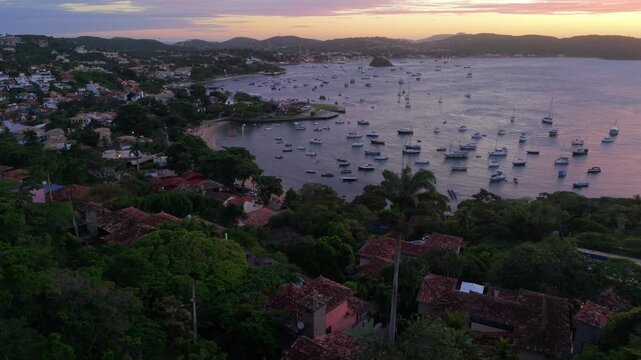 Drone shot captures sunrise over Praia dos Ossos bay, with colorful sky reflected on water, anchored boats, lush greenery, and rustic coastal rooftops in tranquil morning light.