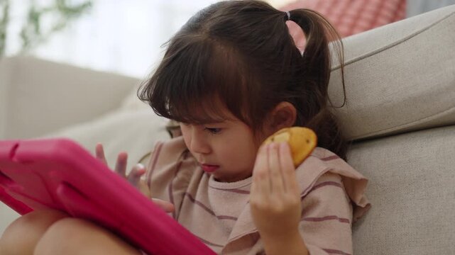 Young girl relaxes on a sofa while watching a pink tablet and holding a small snack at home, illustrating children's screen time, digital entertainment