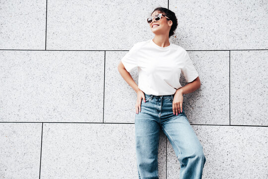 Young beautiful smiling hipster woman in trendy summer white t-shirt and jeans clothes. Carefree woman posing in street. Positive model outdoors near wall. Cheerful and happy. In sunglasses
