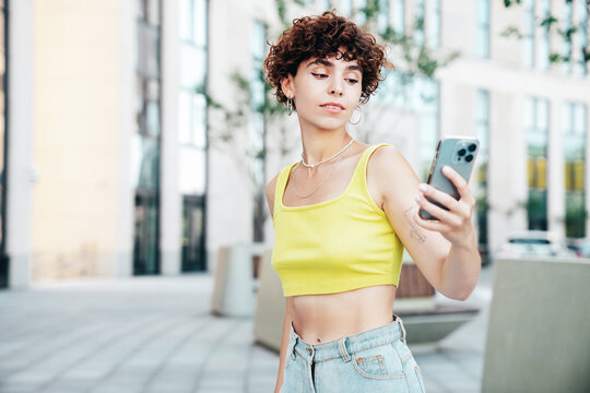Young beautiful smiling hipster woman in trendy summer clothes. Carefree woman with curls hairstyle, in the street. Positive model holds mobile phone, looks at cellphone screen, uses smartphone apps