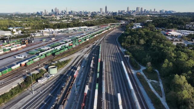 Freight rail yard with stacked intermodal containers, boxcars and parallel tracks stretches toward the downtown Atlanta skyline in Georgia on a clear day. Aerial drone wide shot