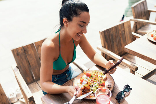 Pretty woman wearing a bikini using her smartphone while drinking a red juice on a terrace near the beach