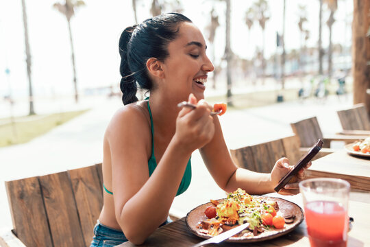 Pretty woman wearing a bikini using her smartphone while drinking a red juice on a terrace near the beach