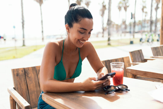 Pretty woman wearing a bikini using her smartphone while drinking a red juice on a terrace near the beach