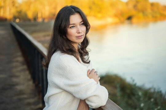Woman in white sweater leaning on railing by river at sunset
