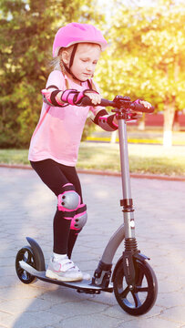 Young girl learns to ride kick scooter on paved path outdoors in sunny park, wearing protective pink helmet and safety gear, a vibrant scene of childhood activity vertical photo