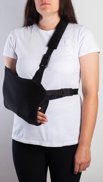 Young adult woman wearing black arm sling for injury support and recovery, dressed in casual white t-shirt and dark leggings, standing against plain background, vertical photo, arm fracture