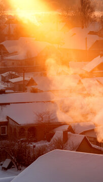 Village rooftops covered by deep snow at sunrise, golden light casts warm glow over houses and rising smoke from chimneys, cold weather atmosphere vertical photo