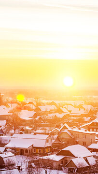 Winter village landscape at golden hour with sun low in horizon, rooftops covered in snow, bare trees visible in tranquil rural setting, vertical photo