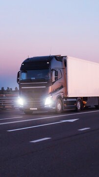 Black semi-truck with white cargo trailer driving on highway during twilight, headlights illuminating road, representing long distance freight transport vertical photo